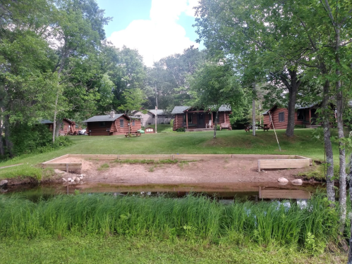 A serene scene of log cabins surrounded by trees, with a small pond and grassy area in the foreground.