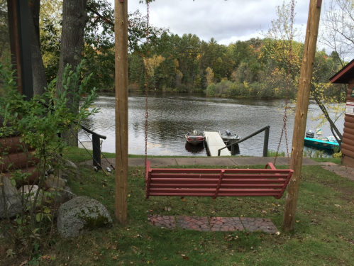 A wooden swing overlooks a calm river with boats docked nearby, surrounded by trees in autumn colors.