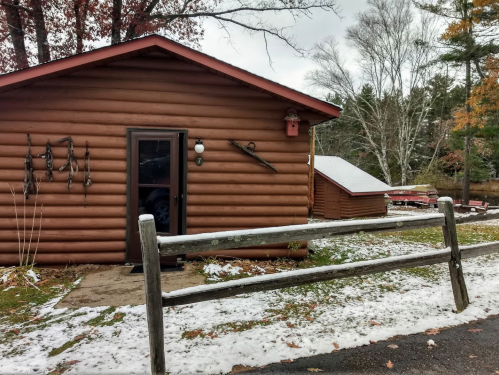 A rustic log cabin with a wooden fence, surrounded by autumn trees and a light dusting of snow.