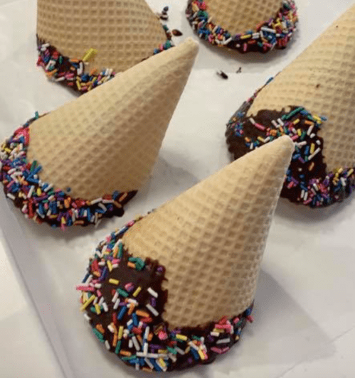 A close-up of ice cream cones dipped in chocolate and covered with colorful sprinkles, arranged on a white surface.