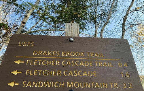 Trail sign indicating directions to Drakes Brook Trail, Fletcher Cascade Trail, and Sandwich Mountain Trail.