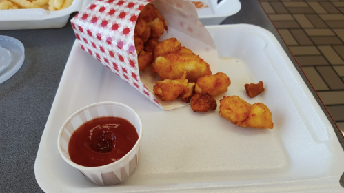 A paper cone filled with golden fried bites next to a small cup of ketchup on a white takeout tray.