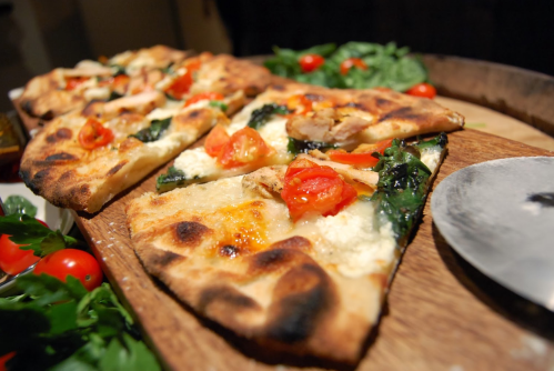 A wooden platter with sliced pizza topped with spinach, tomatoes, and chicken, surrounded by fresh herbs and cherry tomatoes.