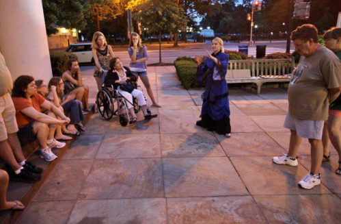 A group of people gathers outdoors, listening to a woman in a blue dress speaking, with a wheelchair user in the foreground.