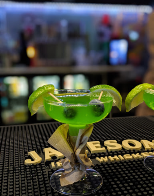 A vibrant green cocktail garnished with lime slices and blueberries, served in a margarita glass on a bar counter.