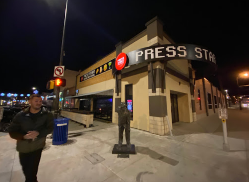 A man walks past a building with a "PRESS START" sign and a statue outside, illuminated at night.