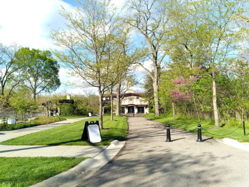 A scenic pathway lined with trees leads to a building, with green grass and a clear blue sky in the background.