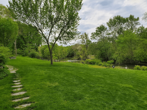 Lush green lawn with a winding path, trees, and a river in the background under a partly cloudy sky.