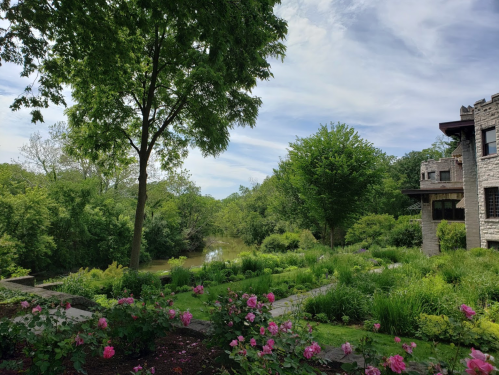 Lush garden with pink flowers, trees, and a serene river view under a partly cloudy sky.