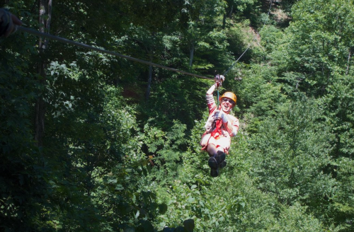 A person in a helmet zip-lining through a lush green forest, smiling with excitement.