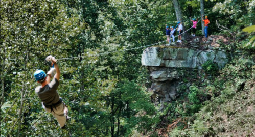 A person zip-lining from a rocky cliff while others watch from a nearby ledge in a forested area.