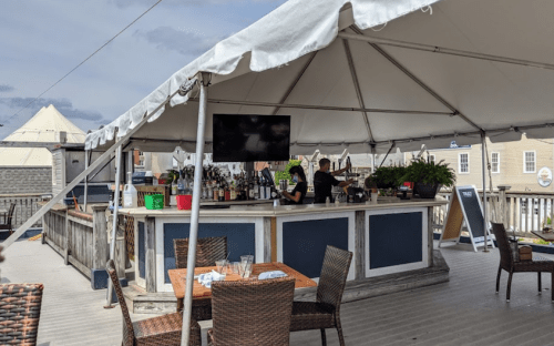 A rooftop bar under a tent with a bartender, tables, and a large TV in a casual outdoor setting.