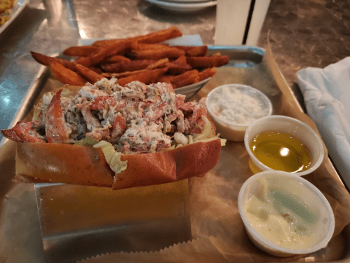 A lobster roll filled with lobster salad on a bun, served with sweet potato fries and two dipping sauces.
