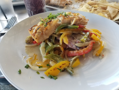 Grilled fish fillet topped with colorful sautéed peppers and onions, served on a white plate with chips in the background.