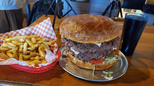 A large burger stacked with patties, lettuce, and tomato, served with crinkle-cut fries and a dark beverage.
