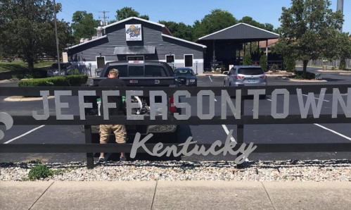 A man stands by a truck in front of a sign that reads "Jeffersontown, Kentucky," with a restaurant in the background.