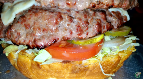 Close-up of a juicy burger with two beef patties, lettuce, tomato, pickles, and mayonnaise on a toasted bun.