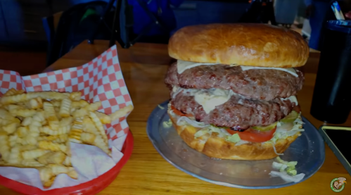 A large burger with two patties, lettuce, and tomato, served with a side of crinkle-cut fries in a red basket.