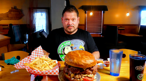 A man sits at a table with a giant burger and fries, looking at the camera in a restaurant setting.