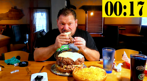 A man is about to eat a large burger, with a timer showing 17 seconds left, in a restaurant setting.