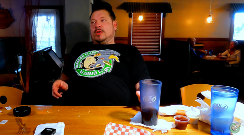 A man in a graphic t-shirt sits at a table with drinks and food remnants, looking thoughtful in a casual dining setting.