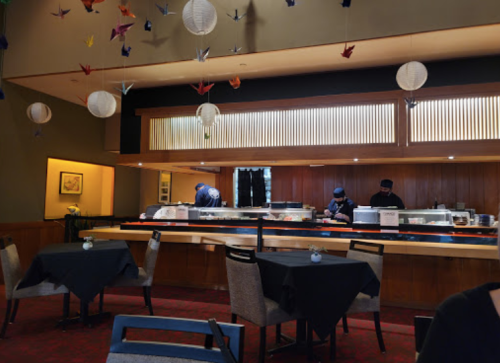 A sushi restaurant interior with black tables, a sushi bar, and chefs preparing food in the background. Colorful paper cranes hang above.