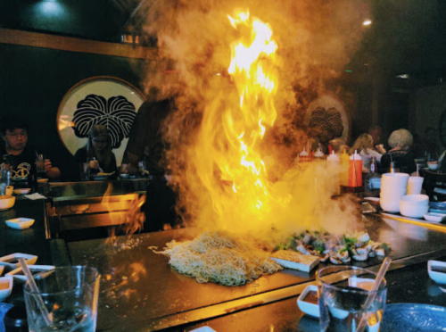 A vibrant flame erupts from a grill at a restaurant, with diners enjoying their meals in the background.