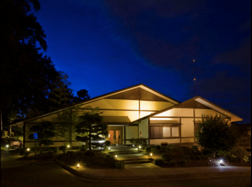 A modern building illuminated at night, surrounded by trees and landscaped gardens under a deep blue sky.
