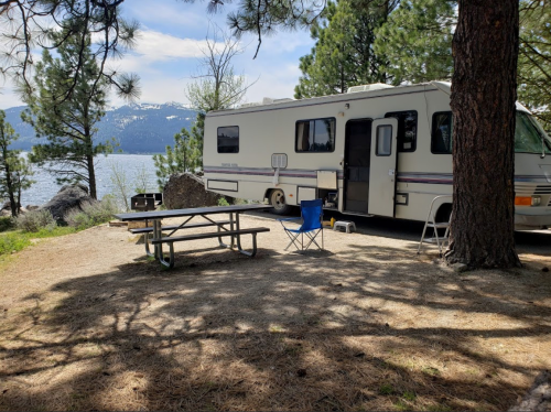 A camper parked by a lake, with a picnic table and chairs under tall trees and mountains in the background.