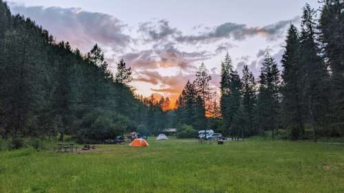 A serene campsite at sunset, featuring tents, picnic tables, and surrounded by tall trees and a colorful sky.