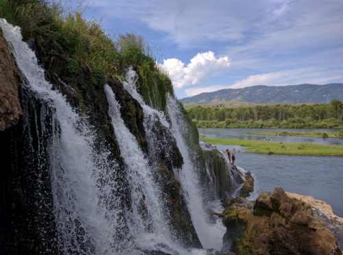 A waterfall cascades over rocks, with two people standing nearby, surrounded by lush greenery and mountains in the background.