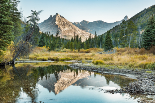 A serene mountain landscape with a calm lake reflecting the peaks and surrounded by lush trees and autumn foliage.