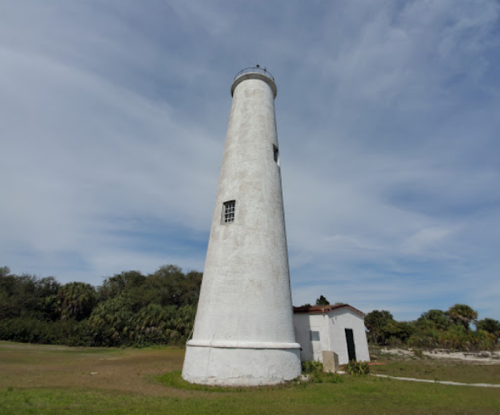 A tall, white lighthouse stands on green grass under a blue sky with scattered clouds.