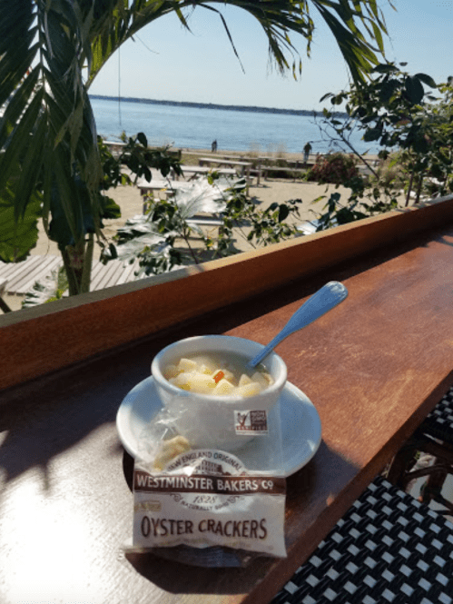 A bowl of clam chowder with oyster crackers on a wooden table, overlooking a scenic waterfront view.