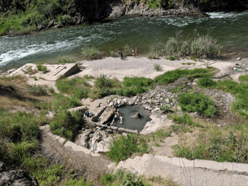 A natural hot spring by a river, with people relaxing in the water and others on the shore, surrounded by greenery.