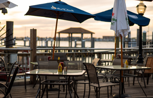 Outdoor dining area with tables, umbrellas, and condiments, overlooking a waterfront at sunset.