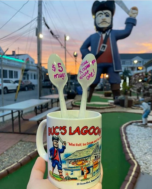 A mug with "Buc's Lagoon" and two spoons, set against a mini-golf course and a large pirate statue at sunset.