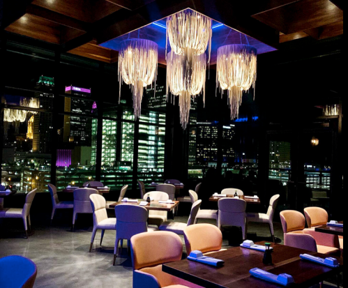A modern restaurant interior at night, featuring stylish lighting and a view of city skyline through large windows.
