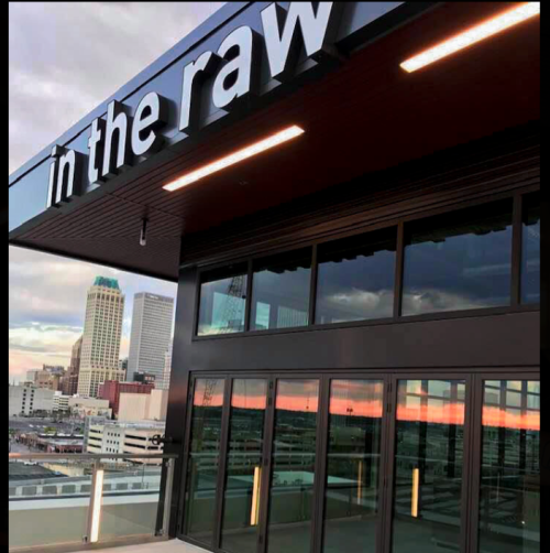 Sign reading "in the raw" on a modern building with city skyline and sunset reflections in the windows.