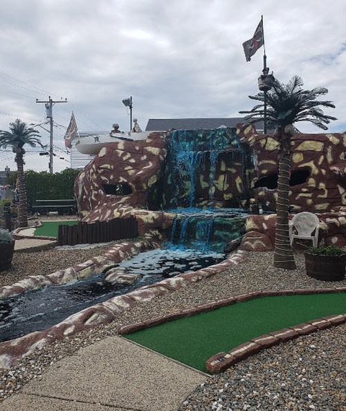 A mini-golf course featuring a waterfall, palm trees, and a rocky landscape under a cloudy sky.