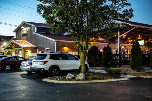 A parking lot with cars near a restaurant, featuring outdoor seating and trees, during twilight.