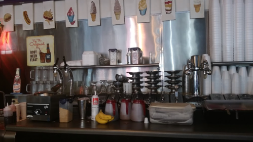 A retro diner counter with ice cream supplies, soda dispensers, and colorful ice cream cone artwork hanging above.