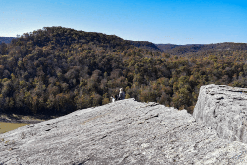 A rocky outcrop overlooks a forested valley with autumn foliage under a clear blue sky.
