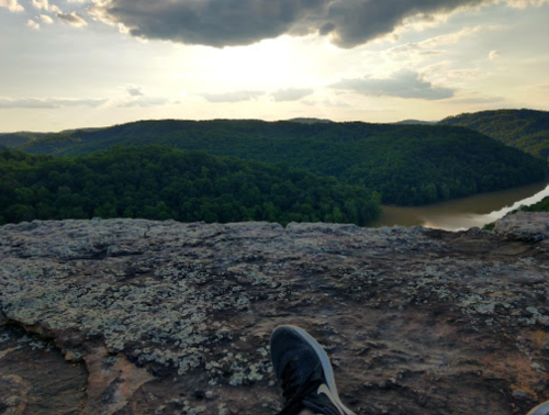 A scenic view from a rocky ledge overlooking a river and lush green hills under a cloudy sky.