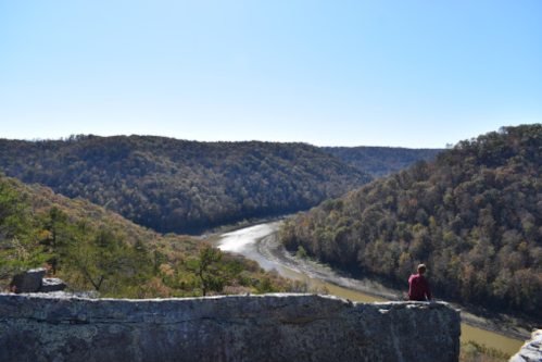 A person sits on a rocky ledge overlooking a winding river surrounded by autumn-colored hills under a clear blue sky.