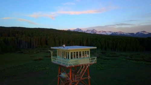 Aerial view of a fire lookout tower surrounded by lush forest and mountains under a colorful sunset sky.