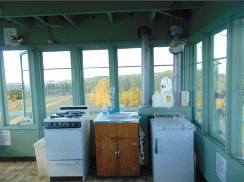 A vintage kitchen with a stove, refrigerator, and wooden cabinet, featuring large windows with a scenic view outside.