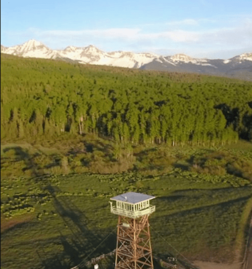 Aerial view of a lookout tower surrounded by lush green trees and mountains in the background under a clear sky.