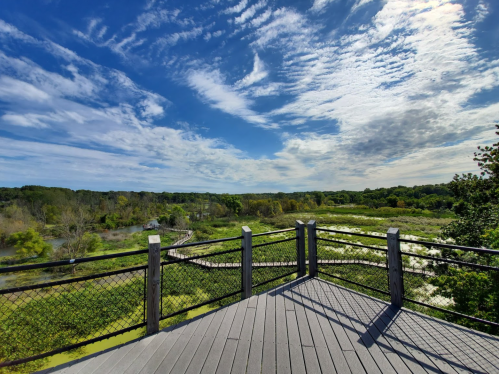 A scenic view from a wooden observation deck overlooking a lush landscape and blue sky with scattered clouds.
