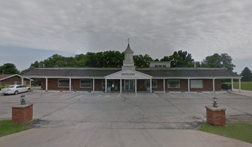 A brick distillery building with a steeple, surrounded by a parking lot and trees under a cloudy sky.